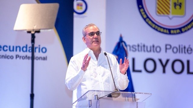El presidente Luis Abinader, con camisa blanca y lentes, habla desde un atril transparente ante un fondo con el nombre del Instituto Politécnico Loyola.