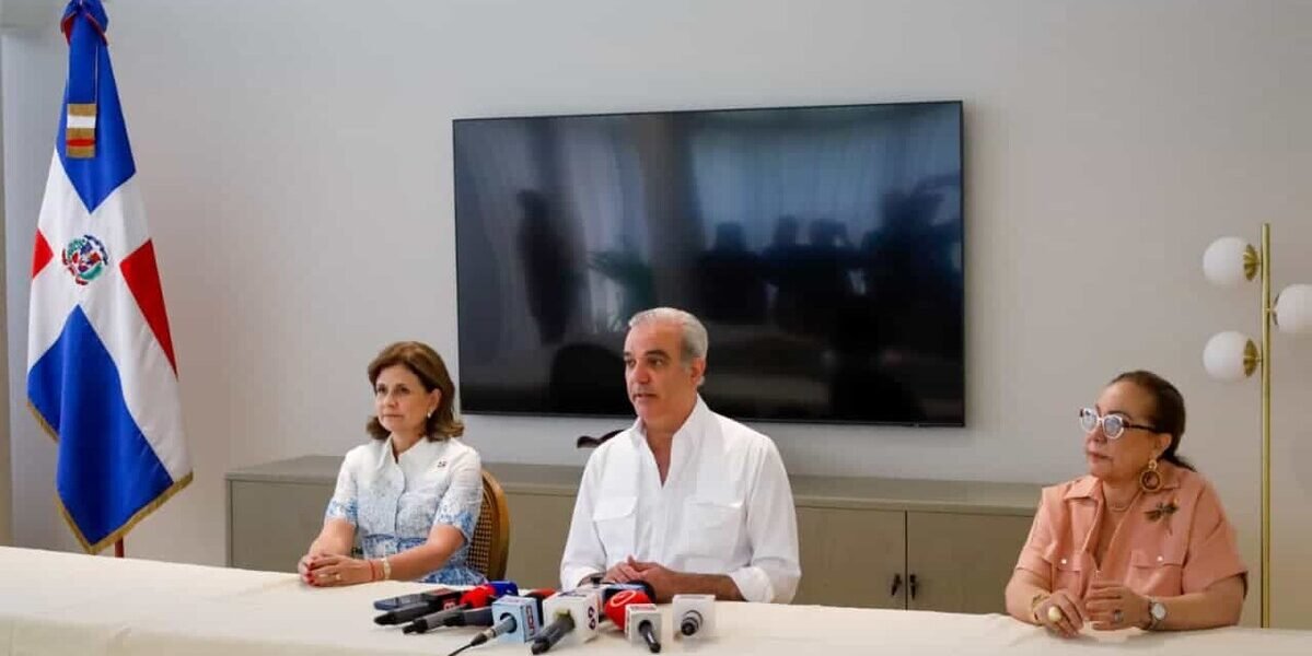 Raquel Peña y Luis Abinader, sentados a una mesa con micrófonos junto a otra mujer, ofrecen declaraciones con una bandera dominicana al fondo.