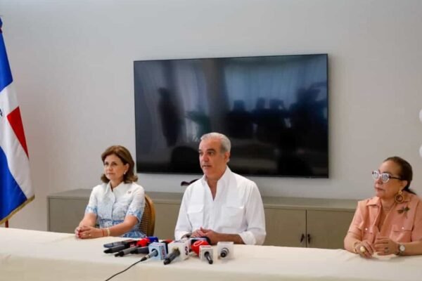 Raquel Peña y Luis Abinader, sentados a una mesa con micrófonos junto a otra mujer, ofrecen declaraciones con una bandera dominicana al fondo.