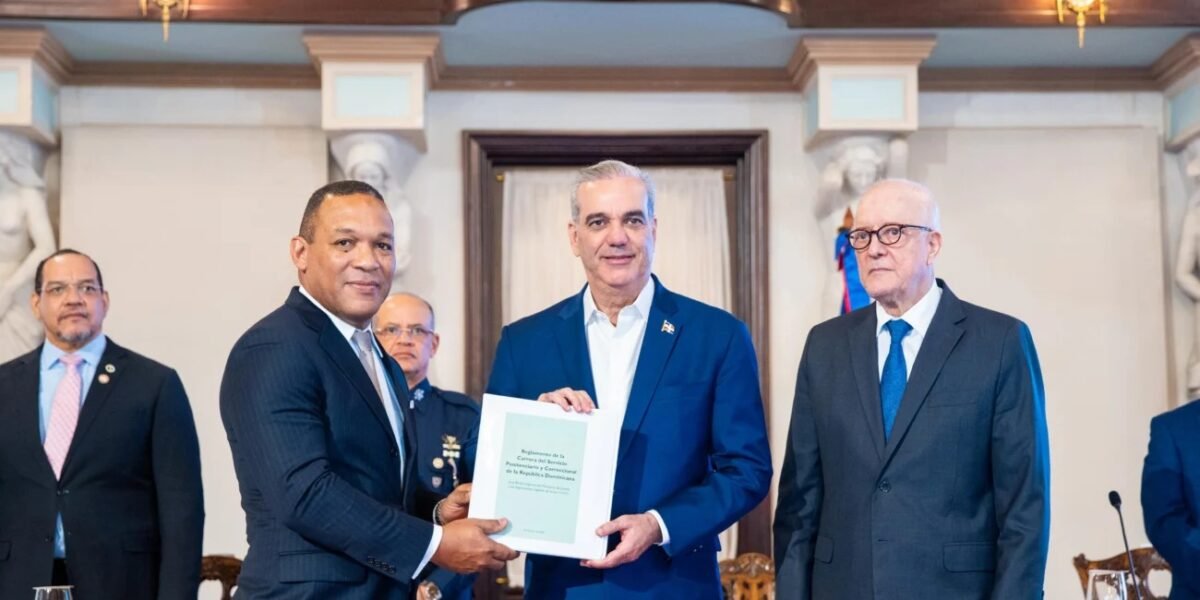El presidente Luis Abinader sostiene junto a otro funcionario un documento titulado Reglamento de la Carrera del Servicio Penitenciario y Correccional de la República Dominicana, en un salón del Palacio Nacional con columnas y esculturas