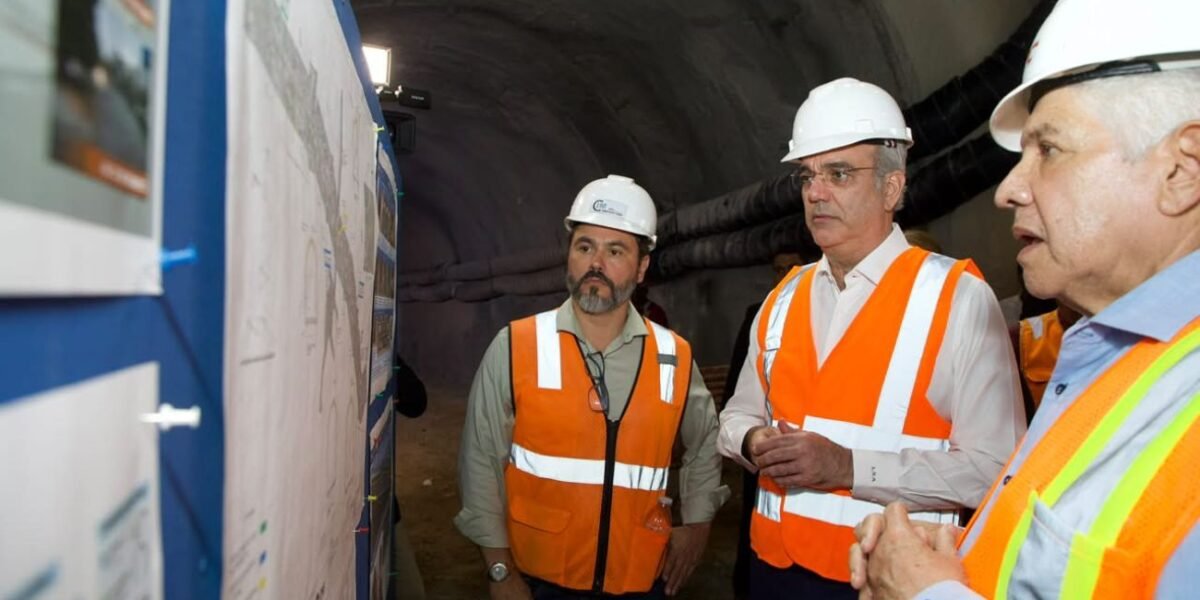 Luis Abinader, con casco blanco y chaleco reflectante, observa planos de obra dentro de un túnel junto a otros dos hombres con equipo de seguridad.