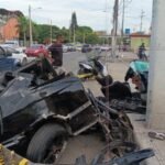 Restos de un carro negro destrozado junto a un poste, acordonados con cinta amarilla, en una avenida con curiosos al fondo