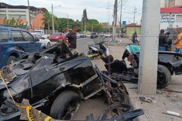 Restos de un carro negro destrozado junto a un poste, acordonados con cinta amarilla, en una avenida con curiosos al fondo