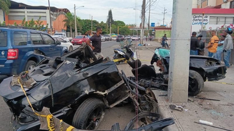 Restos de un carro negro destrozado junto a un poste, acordonados con cinta amarilla, en una avenida con curiosos al fondo