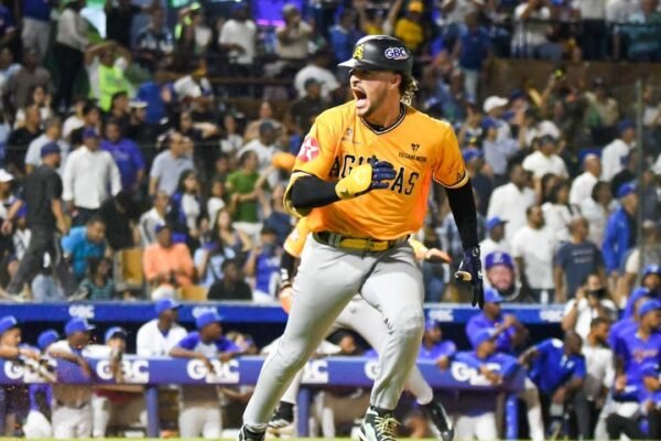 Un jugador de las Águilas Cibaeñas, con camiseta amarilla y casco, grita mientras corre; al fondo, el dugout de los Tigres del Licey y el público en las gradas