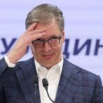 Aleksandar Vučić, con gafas y saco de cuadros, se lleva la mano a la frente ante dos micrófonos frente a un panel con letras en cirílico