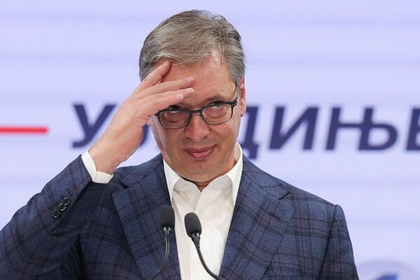 Aleksandar Vučić, con gafas y saco de cuadros, se lleva la mano a la frente ante dos micrófonos frente a un panel con letras en cirílico