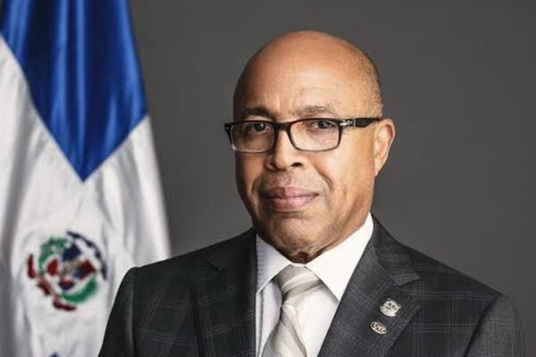 Alfredo Pacheco, con gafas y traje a cuadros, posa junto a la bandera dominicana