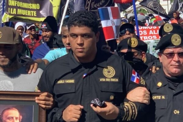 Angelo Vásquez, con uniforme negro de la Antigua Orden Dominicana, marcha tomado del brazo con otros miembros entre banderas dominicanas.