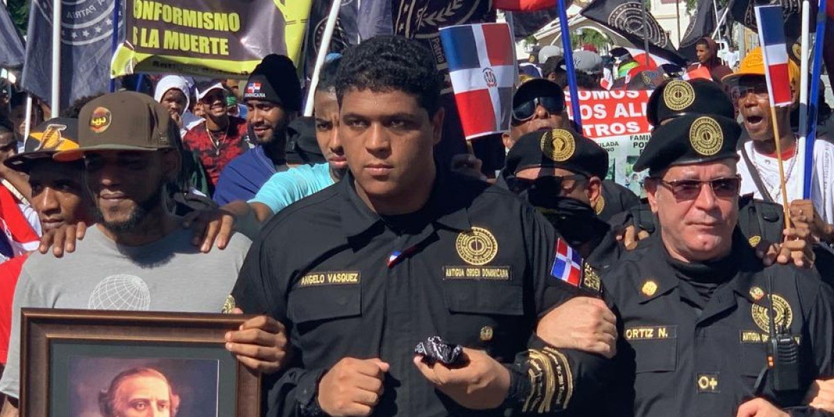 Angelo Vásquez, con uniforme negro de la Antigua Orden Dominicana, marcha tomado del brazo con otros miembros entre banderas dominicanas.