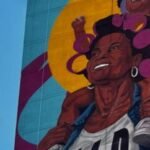 Mural en la fachada de un edificio alto con una mujer sonriente con rolos rosados y una niña sobre sus hombros