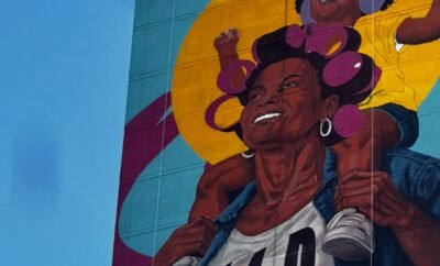 Mural en la fachada de un edificio alto con una mujer sonriente con rolos rosados y una niña sobre sus hombros