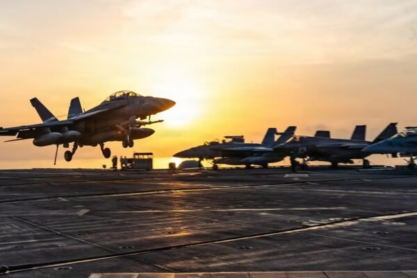 Un avión de combate despega o aterriza en la cubierta de un portaaviones al atardecer, con otros cazas estacionados al fondo.