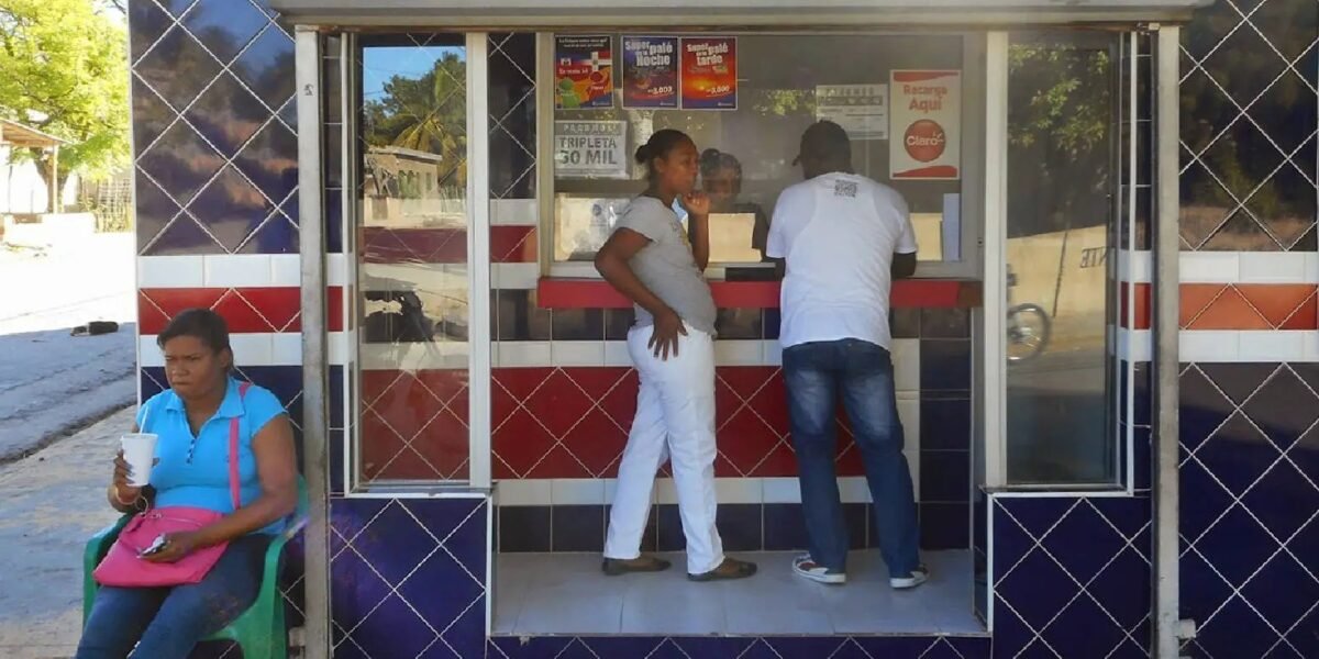 Personas frente a la ventanilla de una banca de lotería con azulejos azules, rojos y blancos, y una mujer sentada en una silla plástica al lado