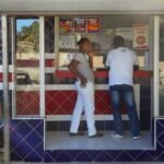 Personas frente a la ventanilla de una banca de lotería con azulejos azules, rojos y blancos, y una mujer sentada en una silla plástica al lado