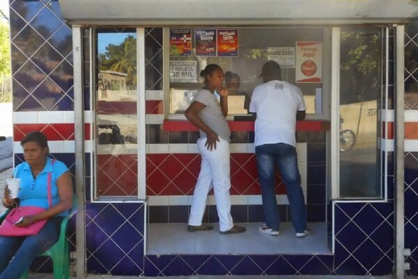 Personas frente a la ventanilla de una banca de lotería con azulejos azules, rojos y blancos, y una mujer sentada en una silla plástica al lado