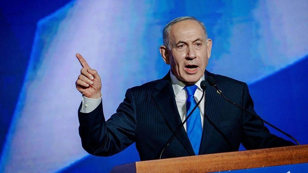 Benjamin Netanyahu, de traje oscuro y corbata azul, habla en un atril con dos micrófonos y señala con el dedo índice, ante un fondo azul