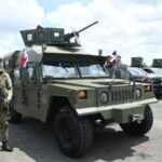 Un militar con boina roja y fusil junto a un vehículo blindado verde con ametralladora y banderas dominicanas; detrás, una fila de camionetas militares
