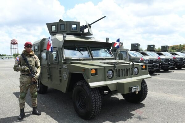 Un militar con boina roja y fusil junto a un vehículo blindado verde con ametralladora y banderas dominicanas; detrás, una fila de camionetas militares