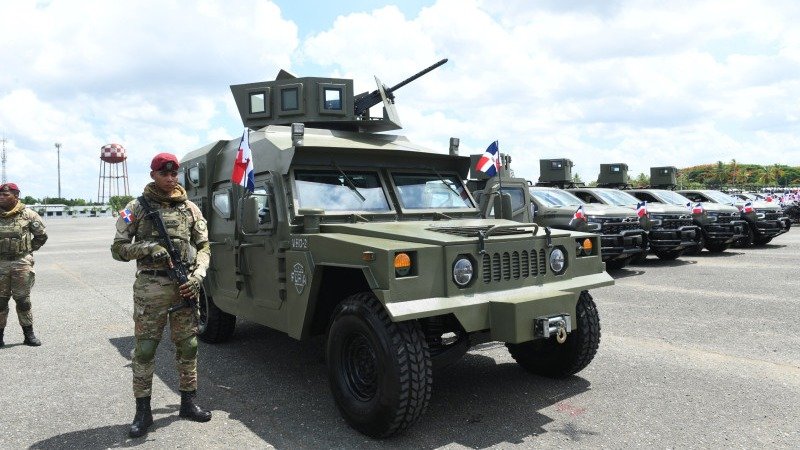 Un militar con boina roja y fusil junto a un vehículo blindado verde con ametralladora y banderas dominicanas; detrás, una fila de camionetas militares