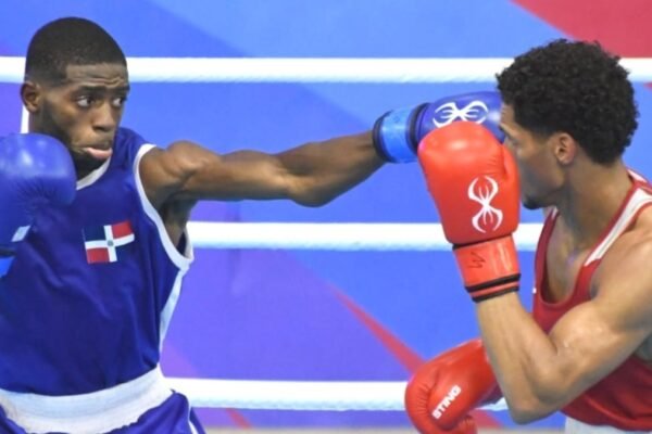 Un boxeador con camiseta azul y la bandera dominicana lanza un golpe a su rival, de rojo, que se cubre con los guantes en el cuadrilátero.