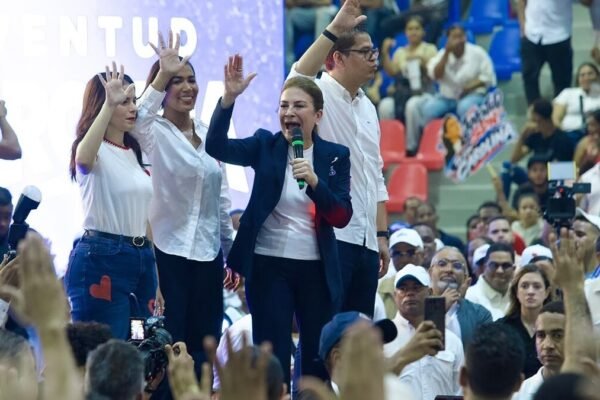 Carolina Mejía, con chaqueta azul y micrófono, levanta la mano en una tarima junto a tres jóvenes, ante un público con las manos alzadas