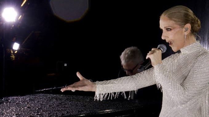 Céline Dion, con vestido plateado de flecos, canta con micrófono y el brazo extendido junto a un piano, de noche