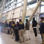 Agentes de la Policía de Investigaciones de Chile, con chaquetas azules que dicen PDI, acompañan a un grupo de personas con maletas en la terminal de un aeropuerto
