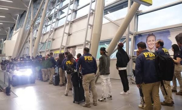 Agentes de la Policía de Investigaciones de Chile, con chaquetas azules que dicen PDI, acompañan a un grupo de personas con maletas en la terminal de un aeropuerto