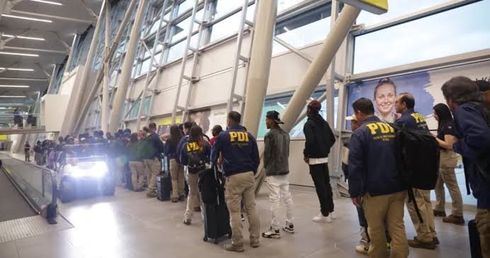 Agentes de la Policía de Investigaciones de Chile, con chaquetas azules que dicen PDI, acompañan a un grupo de personas con maletas en la terminal de un aeropuerto