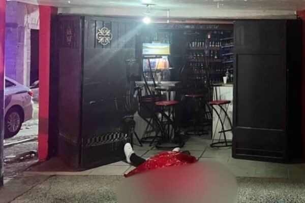 Entrada de un colmado con letrero Los Gozosos, taburetes rojos y una barra; en el suelo, un cuerpo cubierto con una sábana roja, difuminado; al fondo, una patrulla policial