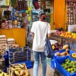 Una mujer de espaldas camina entre cajas de víveres, frutas y verduras frente a un colmado