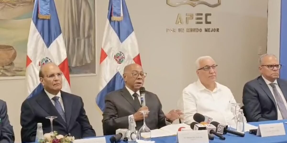 Integrantes de la comisión del Código Penal sentados en una mesa con micrófonos de medios, ante banderas dominicanas y el logo de APEC