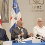 Integrantes de la comisión del Código Penal sentados en una mesa con micrófonos de medios, ante banderas dominicanas y el logo de APEC