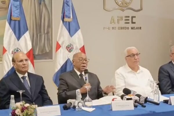 Integrantes de la comisión del Código Penal sentados en una mesa con micrófonos de medios, ante banderas dominicanas y el logo de APEC