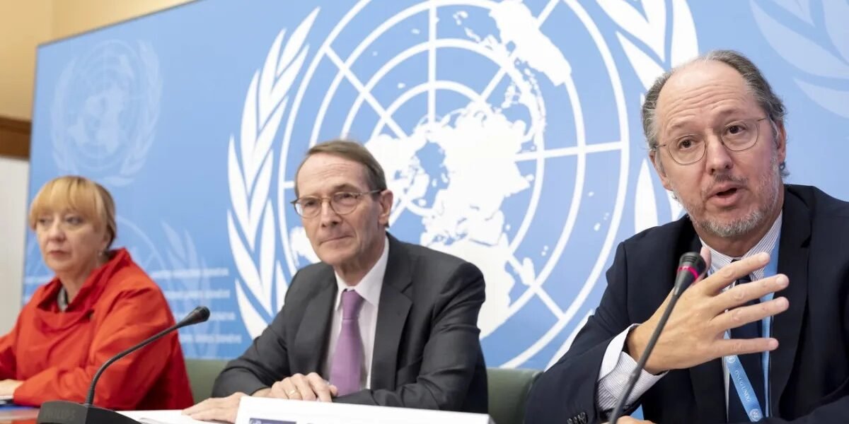 Tres integrantes de una comisión de la ONU sentados ante micrófonos frente al emblema de las Naciones Unidas; a la derecha, un hombre con gafas habla y gesticula detrás de un cartel con el nombre Pablo de Greiff