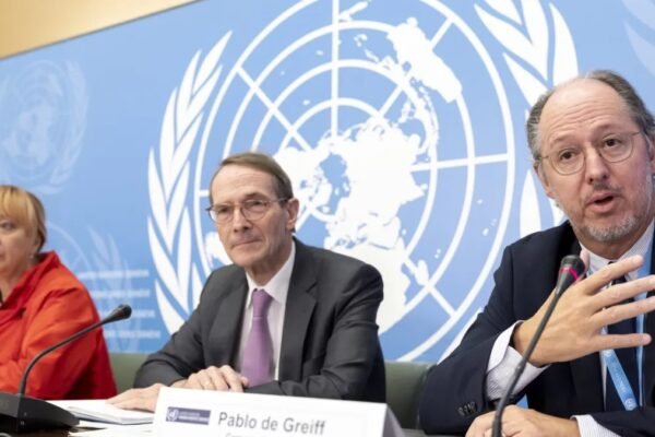Tres integrantes de una comisión de la ONU sentados ante micrófonos frente al emblema de las Naciones Unidas; a la derecha, un hombre con gafas habla y gesticula detrás de un cartel con el nombre Pablo de Greiff