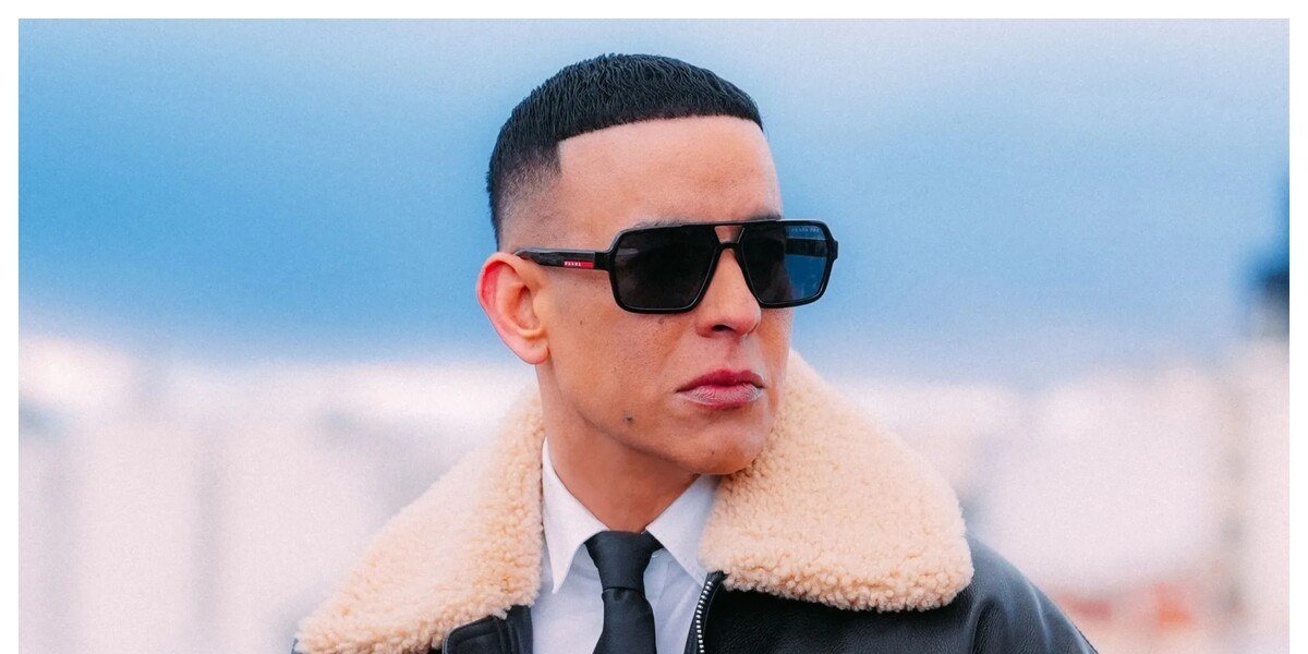 Daddy Yankee con gafas oscuras, camisa blanca, corbata negra y chaqueta de cuero con cuello de borrego, al aire libre.