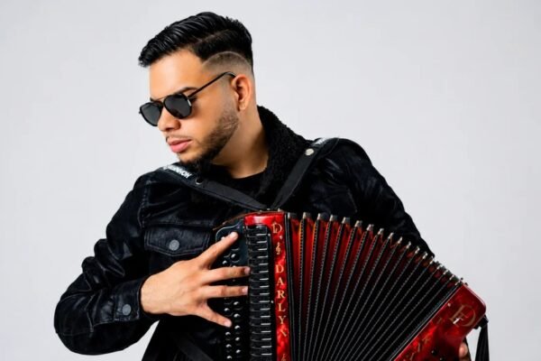 Darlyn Fernández, con gafas oscuras y chaqueta negra, toca un acordeón rojo con su nombre ante un fondo gris claro
