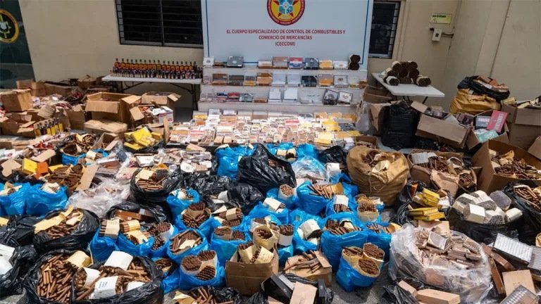 Puros en sacos y cajas, botellas de mamajuana y otras mercancías decomisadas expuestas en el patio del Ceccom, frente a un letrero de la institución