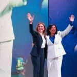 Delia Ortiz, vestida de blanco, y Carolina Mejía, con traje oscuro, saludan con la mano en alto sobre una tarima, frente a una pantalla con el nombre de Delia Ortiz