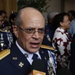 Un oficial de la Policía Nacional con uniforme de gala, lentes y charreteras doradas habla frente a micrófonos de la prensa, rodeado de otros agentes