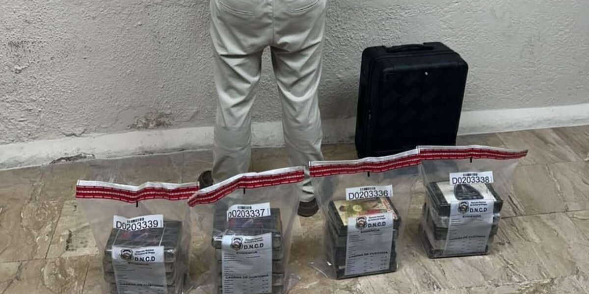 Cuatro bolsas de evidencia de la DNCD con paquetes en su interior, junto a una maleta negra y a una persona de pie de espaldas contra la pared, sin rostro visible