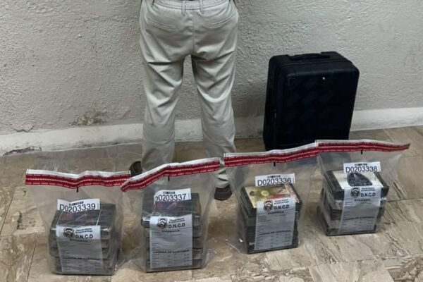 Cuatro bolsas de evidencia de la DNCD con paquetes en su interior, junto a una maleta negra y a una persona de pie de espaldas contra la pared, sin rostro visible