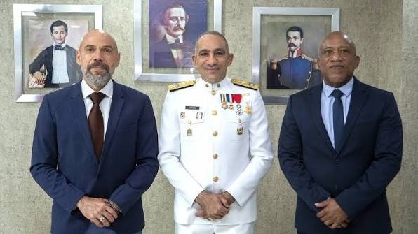 Un oficial con uniforme blanco de gala de la Armada y condecoraciones posa entre dos hombres de traje oscuro, ante tres retratos de próceres