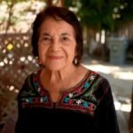 Una mujer mayor de cabello oscuro corto sonríe en un porche con celosías de madera; viste una blusa negra con bordados de flores de colores