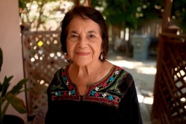 Una mujer mayor de cabello oscuro corto sonríe en un porche con celosías de madera; viste una blusa negra con bordados de flores de colores