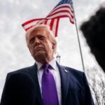 Donald Trump, con corbata morada, frente a micrófonos de la prensa y una bandera de Estados Unidos