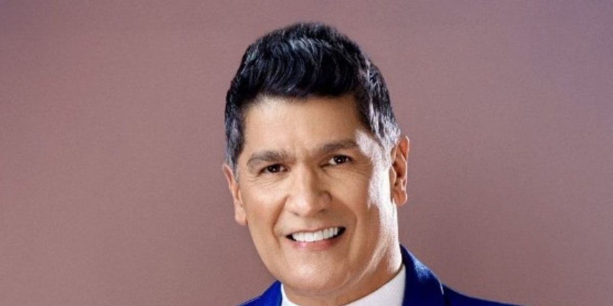 Primer plano de Eddy Herrera sonriente, con chaqueta azul brillante y suéter blanco de cuello alto.