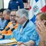 Eduardo Estrella habla por un micrófono sentado a una mesa de reunión junto a otras personas y banderas dominicanas.